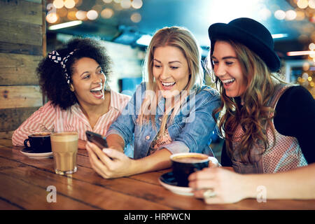 Trois jeunes femmes à une table en bois dans la région de cafe rire, looking at smartphone, quand fille en bleu denim montrant quelque chose sur l'écran pour ses amis Banque D'Images