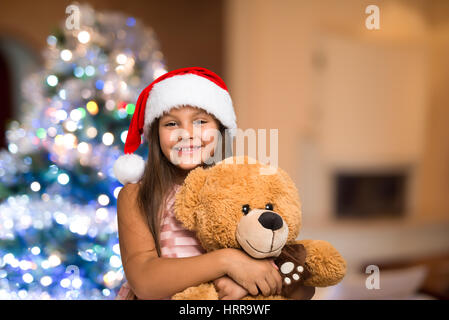 Smiling little girl wearing a Christmas hat holding a teddy bear. Arbre de Noël dans l'arrière-plan Banque D'Images