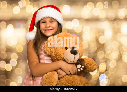 Smiling little girl wearing a Christmas hat holding a teddy bear. Les lumières dans l'arrière-plan Banque D'Images