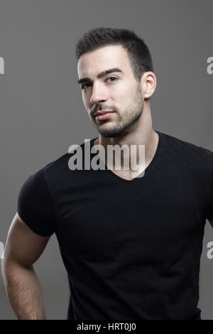Certain macho athletic homme portant un t-shirt noir looking at camera soupçonneusement sur fond gris foncé studio. Banque D'Images