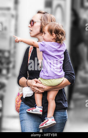 Maman marche sur la rue et porte le jeune enfant qui indique quelque chose qu'elle a remarqué Banque D'Images