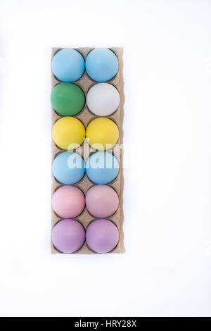 Un oeuf de poule blanche et 11 oeufs de Pâques teints lilas, rose, bleu, vert et jaune dans un carton du dessus avec fond blanc, Copy space Banque D'Images