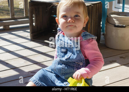 Un an smiling Caucasian baby girl en salopette assis sur une terrasse extérieure avec un verre tasse. New York, USA. Banque D'Images