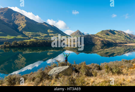 Randonneur debout sur des rochers en tendant les bras, les montagnes se reflétant dans le lac, Moke Lake près de Queenstown, Otago Southland, Région Banque D'Images