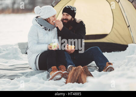 Couple heureux dans des vêtements chauds sont assis dans la neige sur l'arrière-plan d'une tente et un feu brûlant, les amateurs de rire et de profiter du repos et de la s Banque D'Images