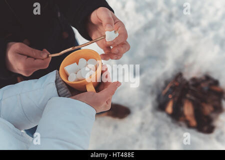 Close up la femme et l'homme les mains au-dessus de la neige et du bois de chauffage. Girl détient tasse avec dessert. Guy est de préparer des aliments pour la cuisson à l'jeu. Le camping d'hiver ou de la randonnée Banque D'Images