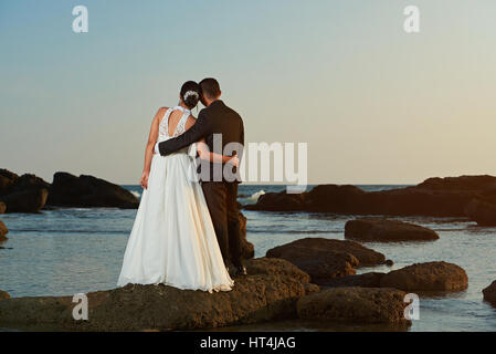 Couple marié regardant le coucher du soleil sur l'ocean beach vue depuis l'arrière. Groom et bride hugging sur le littoral de l'océan Banque D'Images