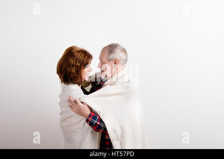 Belle senior couple in love hugging, enveloppé dans une couverture chaude. Studio shot contre le mur blanc. Banque D'Images