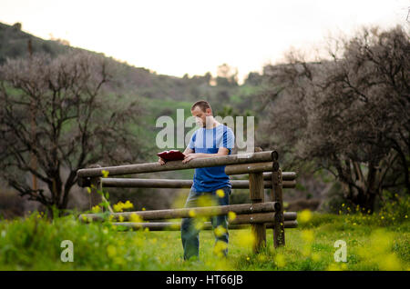 Man reading a book en extérieur dans un domaine appuyé contre une barrière en bois. Banque D'Images