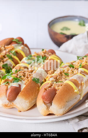 Hot dog avec de la saucisse, oignon frit, des feuilles de coriandre, de moutarde et de la sauce au fromage blanc, servi sur une plaque en céramique avec plus de textile texture béton blanc Banque D'Images