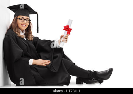 Heureux étudiant diplômé avec un diplôme assis sur le plancher et appuyé contre un mur isolé sur fond blanc Banque D'Images