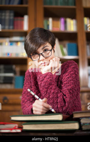 Jeune étudiante avec visage fatigué expression looking at camera avec beaucoup de livres autour de la bibliothèque Banque D'Images