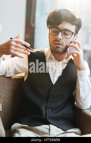 Portrait de barbu aux cheveux noirs concentrés dans des verres en chef et gilet discutent de nouvelles stratégies d'entreprise sur le téléphone et tout en gesticulant hav Banque D'Images