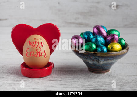 Joyeuses Pâques 2017 le lettrage sur œuf avec support en forme coeur rouge et des œufs en chocolat enveloppé dans du papier d'aluminium Banque D'Images