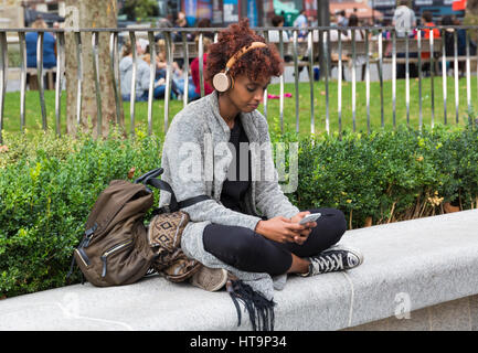 Femme assise sur le mur avec des écouteurs sur looking at phone à Leicester Square, Londres en septembre Banque D'Images