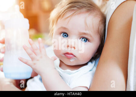 Un petit bébé garçon sur ses genoux de la mère s'est à la recherche à l'appareil photo avec de grands yeux bleus et moustache laiteuse, tenant une bouteille avec sucette. Banque D'Images