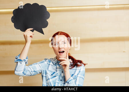 Jolie femme rousse en chemise en jean noir holding speech bubble signer dans la main et de penser à quelque chose contre la paroi en bois Banque D'Images