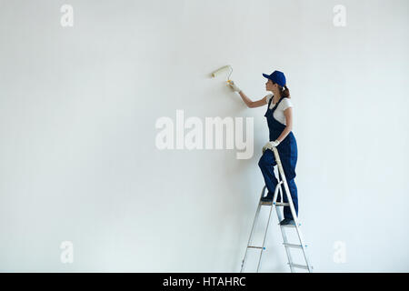 Chambre-peintre en uniforme debout sur l'échelle et la peinture avec rouleau mur Banque D'Images