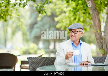 Portrait of senior man wearing cap animé et noir lunettes rim profiter de tasse de café tout en travaillant avec les ordinateurs portables à table de café en plein air sur br Banque D'Images