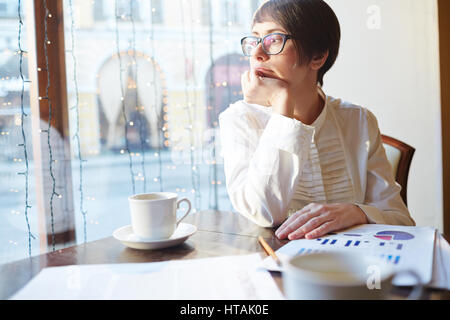 Portrait of pensive young businesswoman wearing white shirt et lunettes à la voiture à la fenêtre assis à table dans un café en soirée Banque D'Images