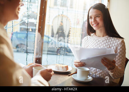 Portrait of young smiling woman hocher à travers les documents pendant les négociations réunion au cafe table en soirée Banque D'Images