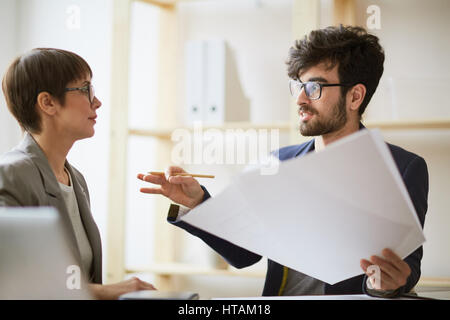 Portrait de l'homme créatif barbu holding tas du papier des gestes tout en expliquant les détails du projet activement de businesswoman assise à côté de lui en mod Banque D'Images