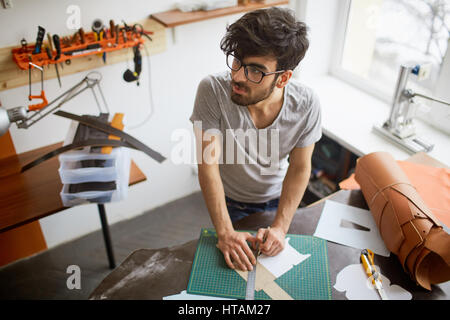 High angle portrait of middle eastern musculaire homme portant des lunettes et coupe de créatif à l'écart, songeur, tout en créant des dessins et modèles industriels en cuir w Banque D'Images