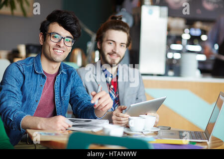 Portrait de deux jeunes hommes créatifs habillés en tenue d'affaires décontractée smiling at camera while working at table lors de réunion Banque D'Images