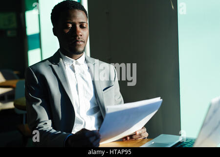 Portrait d'Afro-Américain businessman sitting in sunlight at desk in office et la lecture des documents avec l'expression grave Banque D'Images