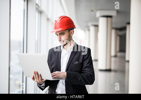 Beau mâle ingénieur est à l'aide d'un ordinateur portable pour travailler. L'homme est un casque blanc est à la recherche à l'écran avec l'aspiration. Banque D'Images