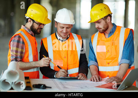 La construction de l'équipe de travailleurs portant des casques de protection et de gilets pour discuter des détails du projet avec permanent superviseur exécutif à table avec blueprin Banque D'Images