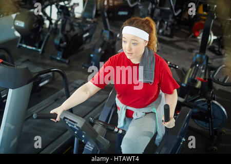 High angle portrait of cute surpoids femme aux cheveux rouges la formation en utilisant les machines elliptiques in gym Banque D'Images