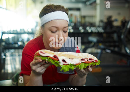 Portrait of cute surpoids femme léchant ses lèvres voulant manger énorme sandwich d'engraissement pendant l'entraînement en salle de sport, luttant pour rester en forme Banque D'Images