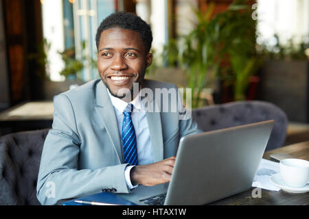 Portrait of handsome African- American businessman wearing costume formel en souriant gaiement tout en travaillant avec laptop in cafe Banque D'Images