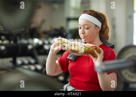 Portrait de jeune femme en surpoids de lécher ses lèvres à propos de manger gros sandwich engraissement prendre une pause pour s'entraîner dans la salle de sport, concept de l'obsession alimentaire Banque D'Images