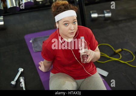 High angle portrait of cute femme obèse faisant des exercices de remise en forme et d'écouter de la musique dans les écouteurs pendant l'entraînement sur le tapis de gym, faire une pause t Banque D'Images