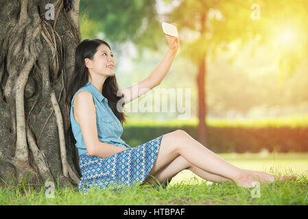 Belle femme asiatique en tenant sitting on grass selfies en parc extérieur. nature dehors arrière-plan. les gens et la technologie concept Banque D'Images