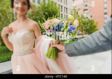 Bride and Groom tenir un bouquet. L'accent sur le bouquet. Couple nouvellement marié à pied sur les chemins se tenant la main et à la recherche à l'autre. À l'arrière du bâtiment Banque D'Images