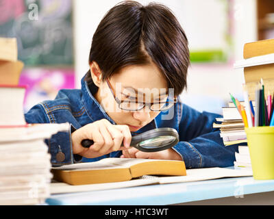 Élève d'Asie avec des lunettes à l'aide d'une loupe pour agrandir les mots dans un livre épais. Banque D'Images