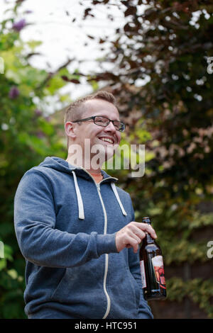 Cheveux blonds courts jeune homme portant des lunettes et capuche bleu potable cavalier ale. Réunion d'amis après le travail, un barbecue dans le jardin. Shot verticale Banque D'Images