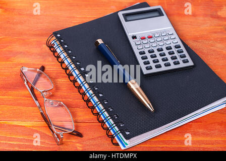 Journal noir, stylo, calculatrice et verres allongé sur une surface en bois. Banque D'Images