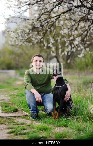 Jeune homme est assis sur l'herbe avec dobermann chien sur un fond d'arbres en fleurs de printemps Banque D'Images