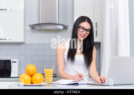 Jolie fille brune dans les verres raboteuse jour, l'étude ou de l'écriture accueil tâche en cuisine en matinée. Avec succès d'étudiants et personnes à l'aide d'ordinateur portable et l'IRG Banque D'Images