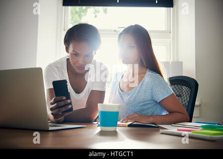 Deux jeunes femmes collègues de travail dans une nouvelle start-up de la vérification d'une petite entreprise téléphone message ensemble à un bureau dans le bureau avec une fenêtre de sun flare beh Banque D'Images