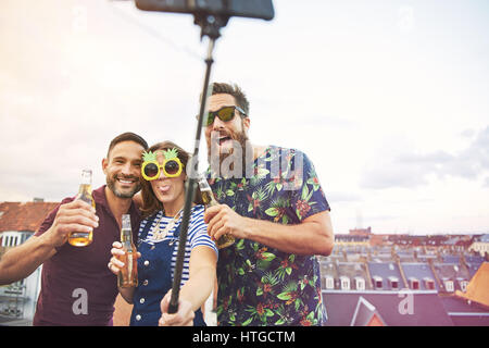 Groupe de trois jeunes adultes ivres d'alcool et de prendre des photos d'eux-mêmes sur le toit avec toit sur stick selfies avec copie espace à ciel clair Banque D'Images