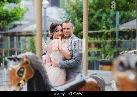 De jeunes mariés heureux à la recherche à l'appareil photo. Heureux nouveaux mariés huging près du carrousel. Les chevaux du carrousel en premier plan. Banque D'Images