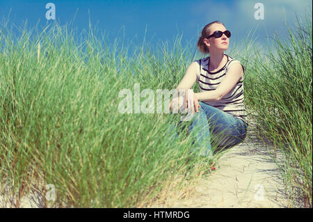 Jeune femme assise dans les dunes de sable entre les hautes herbes, se détendre en profitant de la vue sur la journée ensoleillée, Luskentyre, Isle of Harris, Scotland Banque D'Images