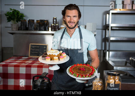 Portrait de personnel masculin holding dessert on cake stand at counter in coffee shop Banque D'Images