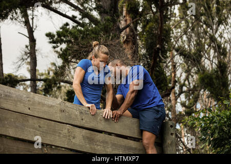 Male trainer assisting woman d'escalader un mur en bois au cours de parcours Banque D'Images