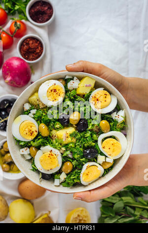 Une femme tenant un bol de salade de pommes de terre de style méditerranéen avec des herbes et oeufs durs accompagné d'olives vertes et noires, oignons rouges, sumac et Banque D'Images